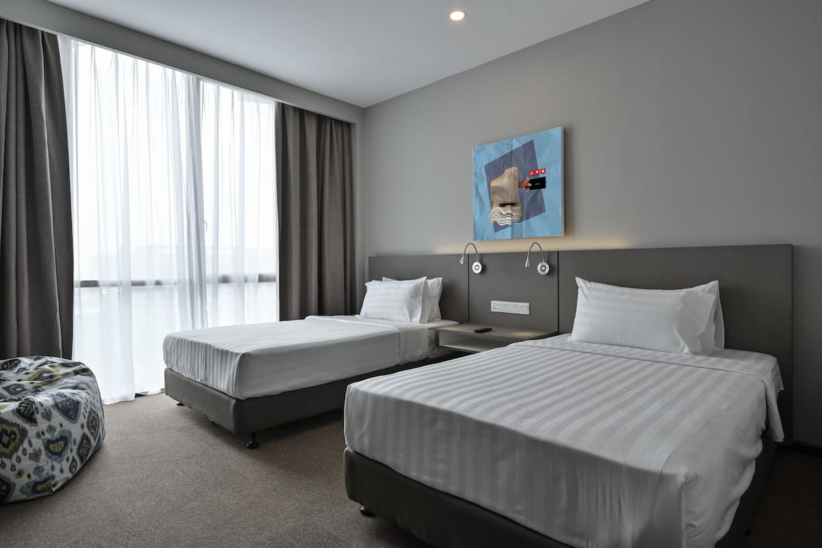 Ein Hotelzimmer mit zwei Betten, einem großen Fenster mit Vorhängen, einem Wandbild und einer kleinen Sitzecke.
