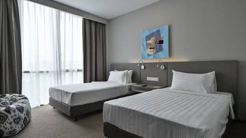 A hotel room with two beds, a large window with curtains, a wall-mounted artwork, and a small seating area.