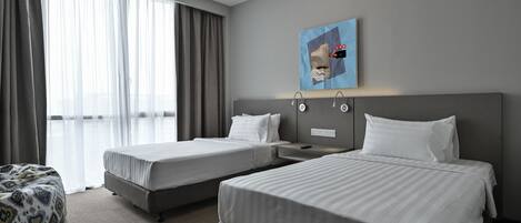 A hotel room with two beds, a large window with curtains, a wall-mounted artwork, and a small seating area.
