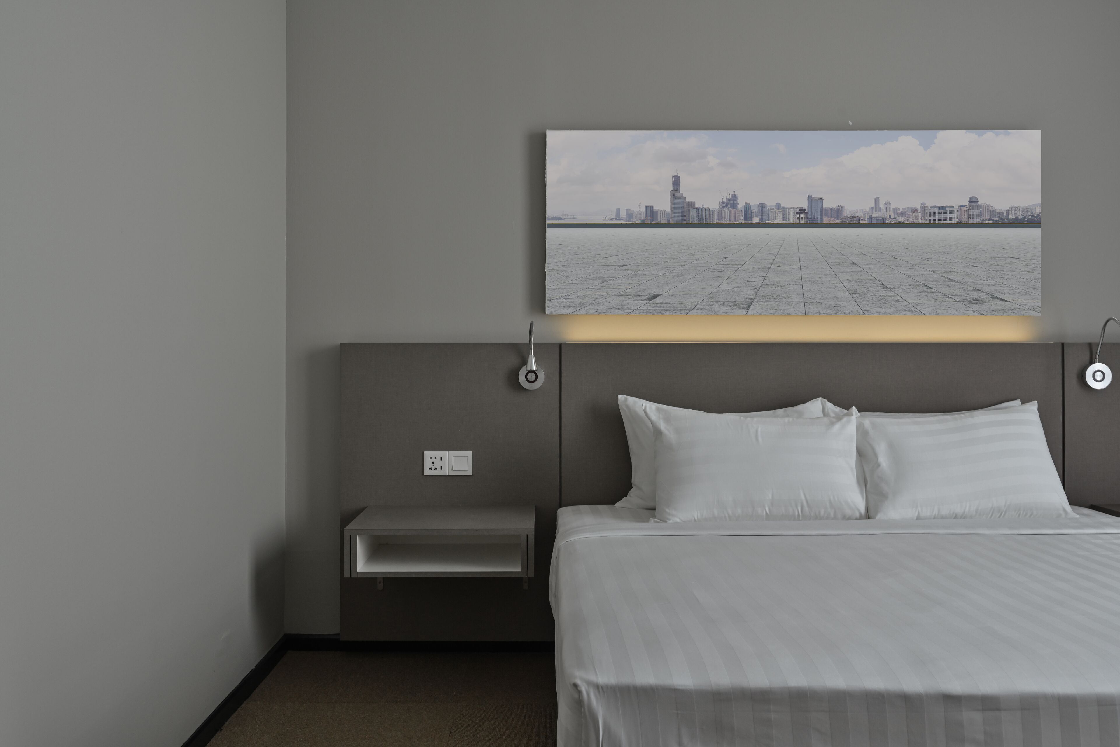 A hotel room with a bed, a bedside table, a wall-mounted lamp, and a cityscape view.