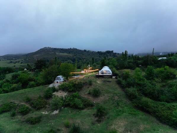 A rural landscape with a large white dome structure surrounded by greenery, a smaller dome, and a path leading to a building with lights. - Glamping Dream Domes Ismayilli (Ismailli)
