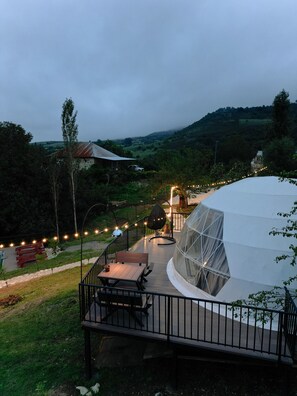 A dome-shaped tent with a transparent roof, a wooden deck with a table and chairs, and string lights along the deck's railing. - Glamping Dream Domes Ismayilli (Ismailli)