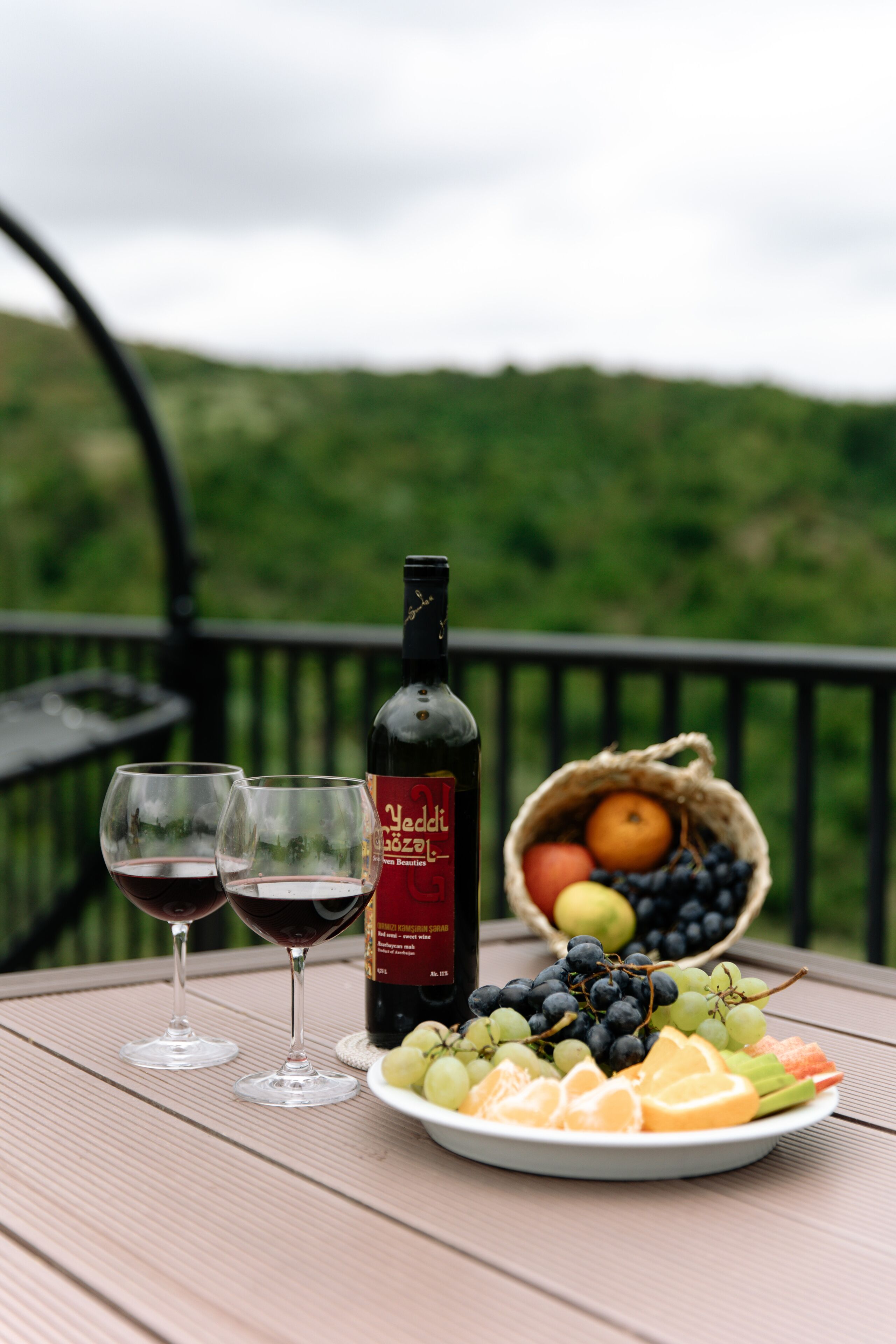 A table with a bottle of wine, two glasses of red wine, a plate of fruit, and a basket of additional fruit.