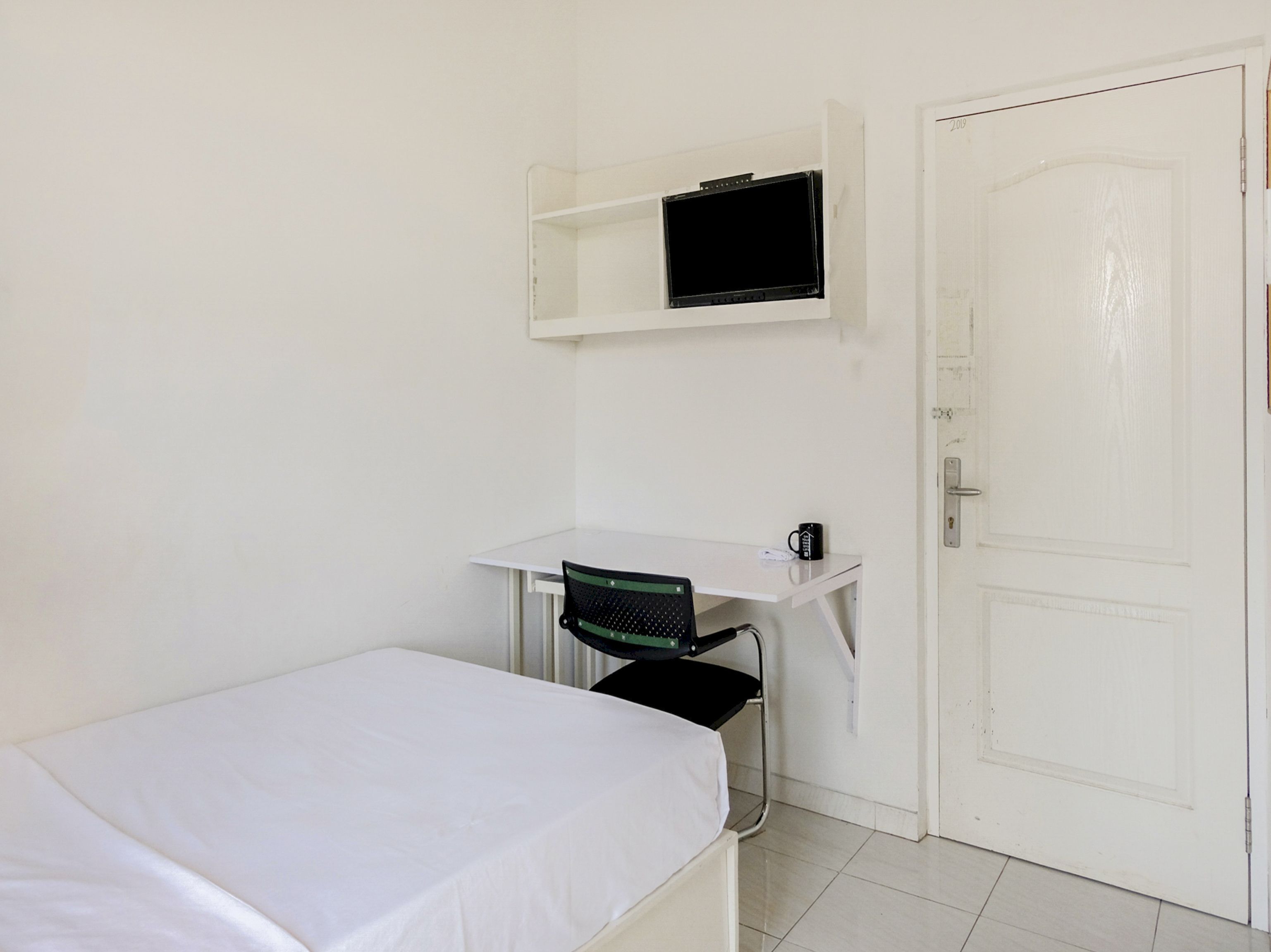 Une petite pièce propre, avec un lit simple, un bureau avec une chaise, un téléviseur fixé au-dessus du bureau et une porte.