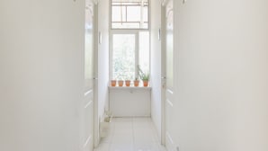 A narrow hallway with white walls, a window with potted plants, and a door on the left. - OYO Life 2760 Kost Simpang Borobudur 47 (Malang)