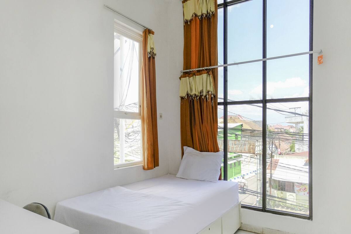 A small, clean room with a single bed, a window with curtains, and a view of buildings outside.
