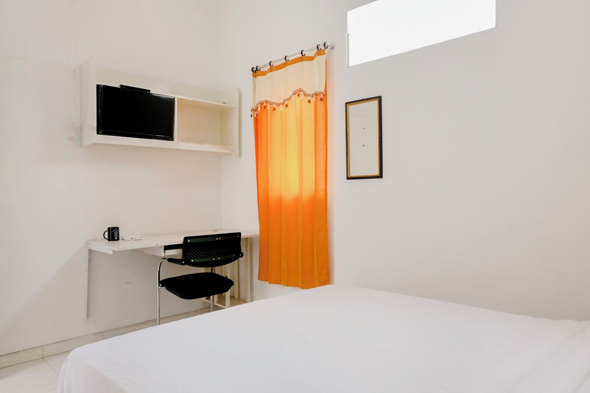 A bedroom with a bed, a desk with a chair, a TV mounted on the wall, and a window with a curtain.