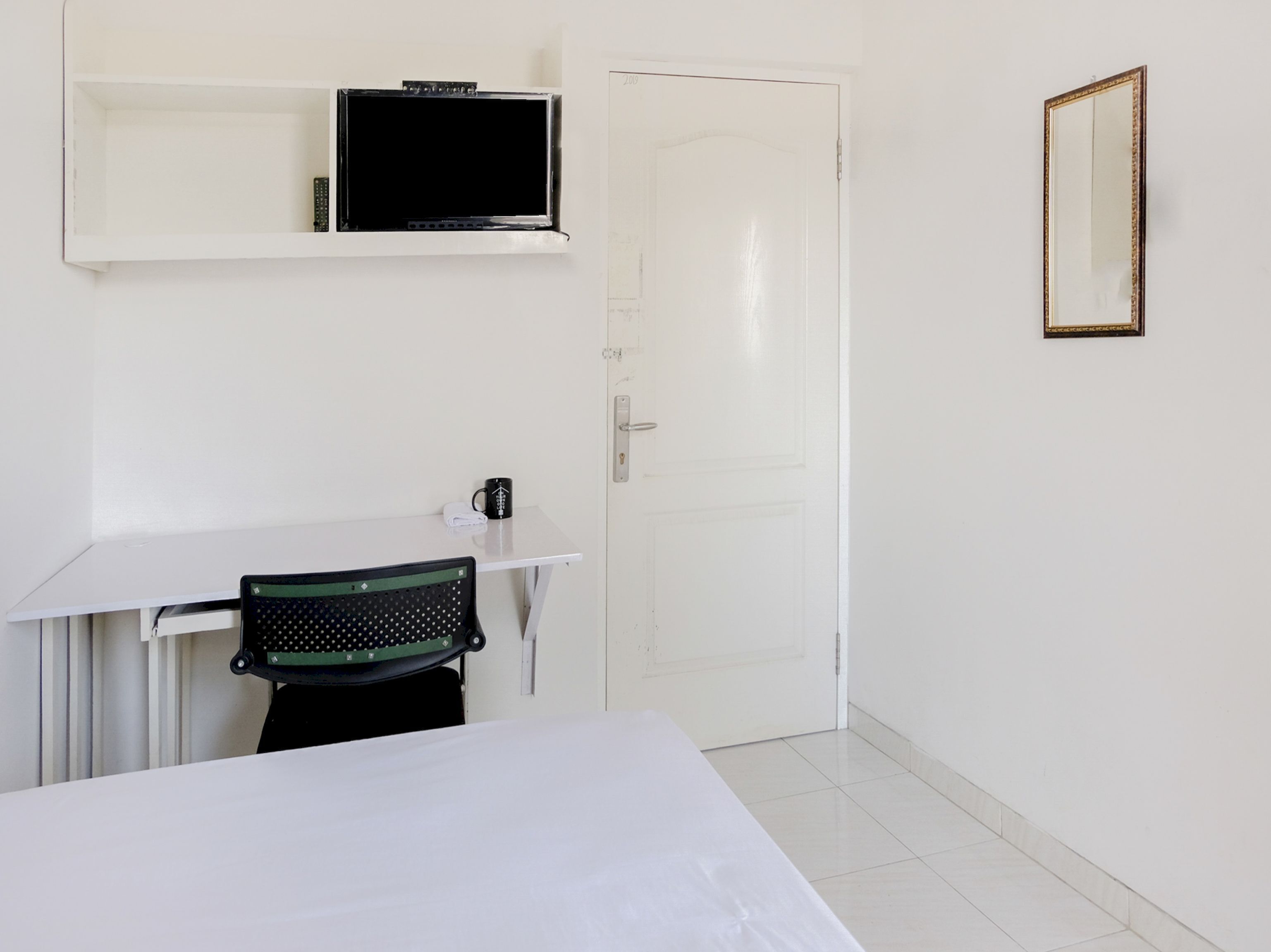 Ein kleines, sauberes Zimmer mit weißem Schreibtisch, Stuhl, Bett, Fernseher auf einem Regal und einer Tür.