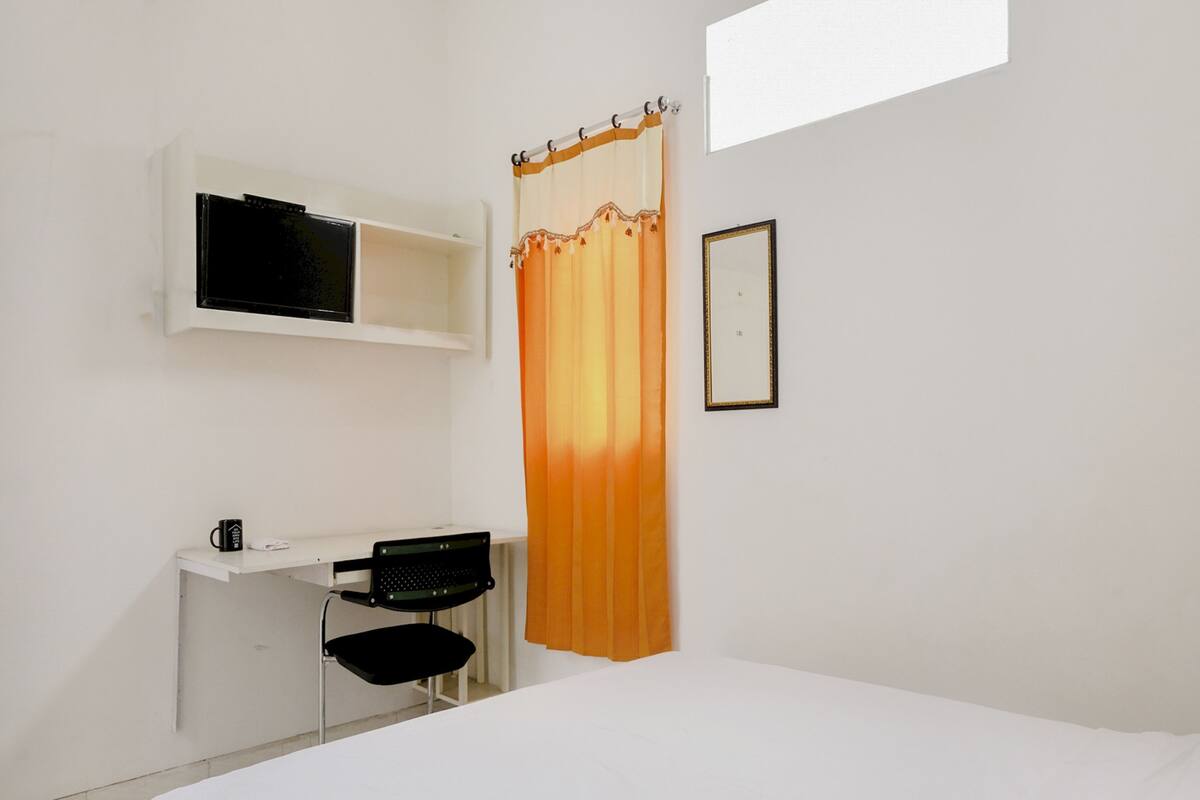 A bedroom with a bed, a desk with a chair, a shelf with a TV, and a curtain.