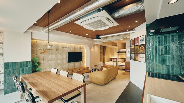 Een moderne hotelkamer met een houten eettafel, stoelen, een bank, een aan de muur bevestigde televisie en een groene betegelde spatwand.