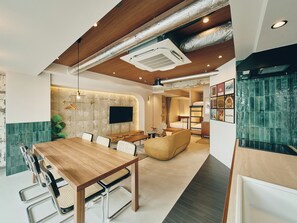 Quarto de hotel moderno com mesa de jantar de madeira, cadeiras, sofá, TV fixada na parede e painel de azulejos verdes.