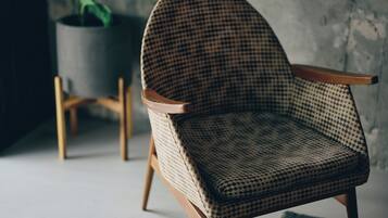 Una silla con estampado de cuadros y reposabrazos de madera, ubicada en una habitación con pared de hormigón y una planta en maceta al fondo.