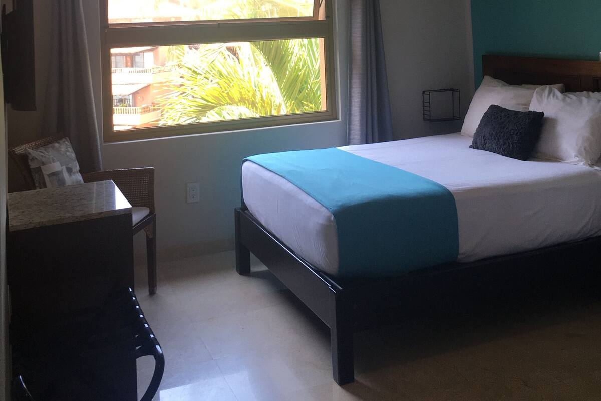 A bedroom with a bed, a desk, a chair, a window with curtains, and an air conditioning unit.