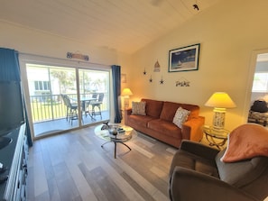 A living room with a sofa, a coffee table, a TV, and a balcony with outdoor furniture.