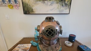 Ein Taucherhelm im Vintage-Stil auf einem Holztisch, ein Buch mit dem Titel 'Willkommen am Strand', eine blaue Tasse und ein Kartenspiel.