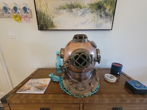 Un casque de plongée de style vintage posé sur une table en bois, un livre intitulé « Bienvenue à la plage », une tasse bleue et un jeu de cartes.