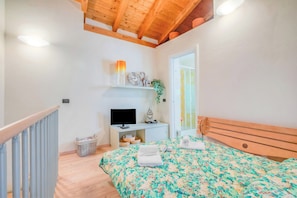 Un dormitorio con techo de madera, cama con colcha floral, televisor sobre un mueble blanco y ventana con vistas.