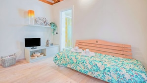 A bedroom with a wooden ceiling, a bed with a floral bedspread, a television on a white stand, a small shelf with decorative items, and a basket on the floor. - La Piccola Casa Dell'uva Fragola Next to the Lake (Meina)