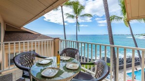 A balcony with a table set for two, overlooking a pool and the ocean. - Sea Village 3312: Oceanfront condo in Kailua-Kona, with stunning views! (Kailua-Kona)