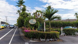 A road sign for "The Sea Village" with lush greenery and palm trees. - Sea Village 3312: Oceanfront condo in Kailua-Kona, with stunning views! (Kailua-Kona)