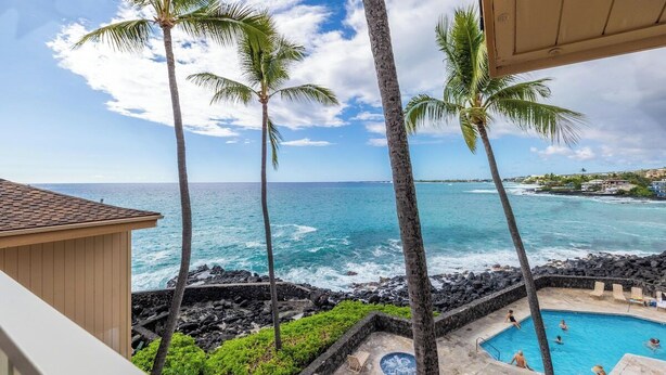 A pool area with a view of the ocean, surrounded by palm trees and a stone wall. - Sea Village 3312: Oceanfront condo in Kailua-Kona, with stunning views! (Kailua-Kona)