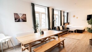 A modern living room with a wooden dining table, a sofa, and a large window with curtains.