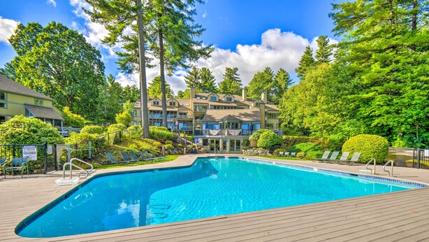 A swimming pool with a wooden deck, surrounded by lush greenery and a multi-story building with balconies. - Gilford Townhome w/ Lakefront Beach & Pool Access! (Gilford)