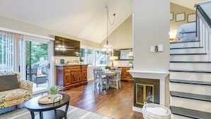 A spacious living area with a sofa, coffee table, and dining table. A kitchen with wooden cabinets and a fireplace is visible in the background. - Gilford Townhome w/ Lakefront Beach & Pool Access! (Gilford)