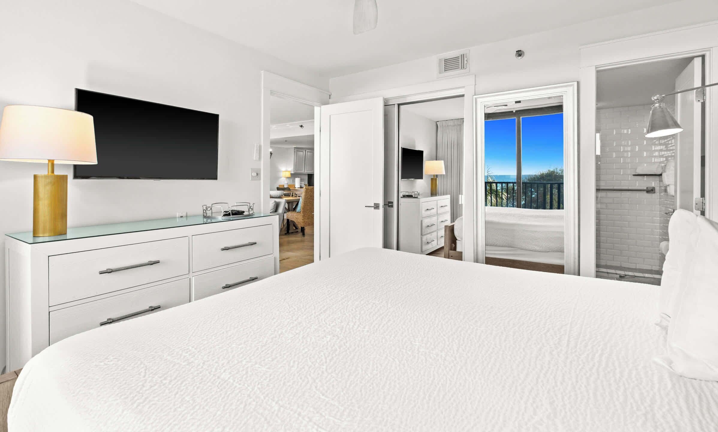 Ein modernes Hotelzimmer mit einem großen Bett, einem flachbildfernseher an der Wand, einem Glastisch mit Lampe sowie einem Badezimmer mit Dusche und Balkon.