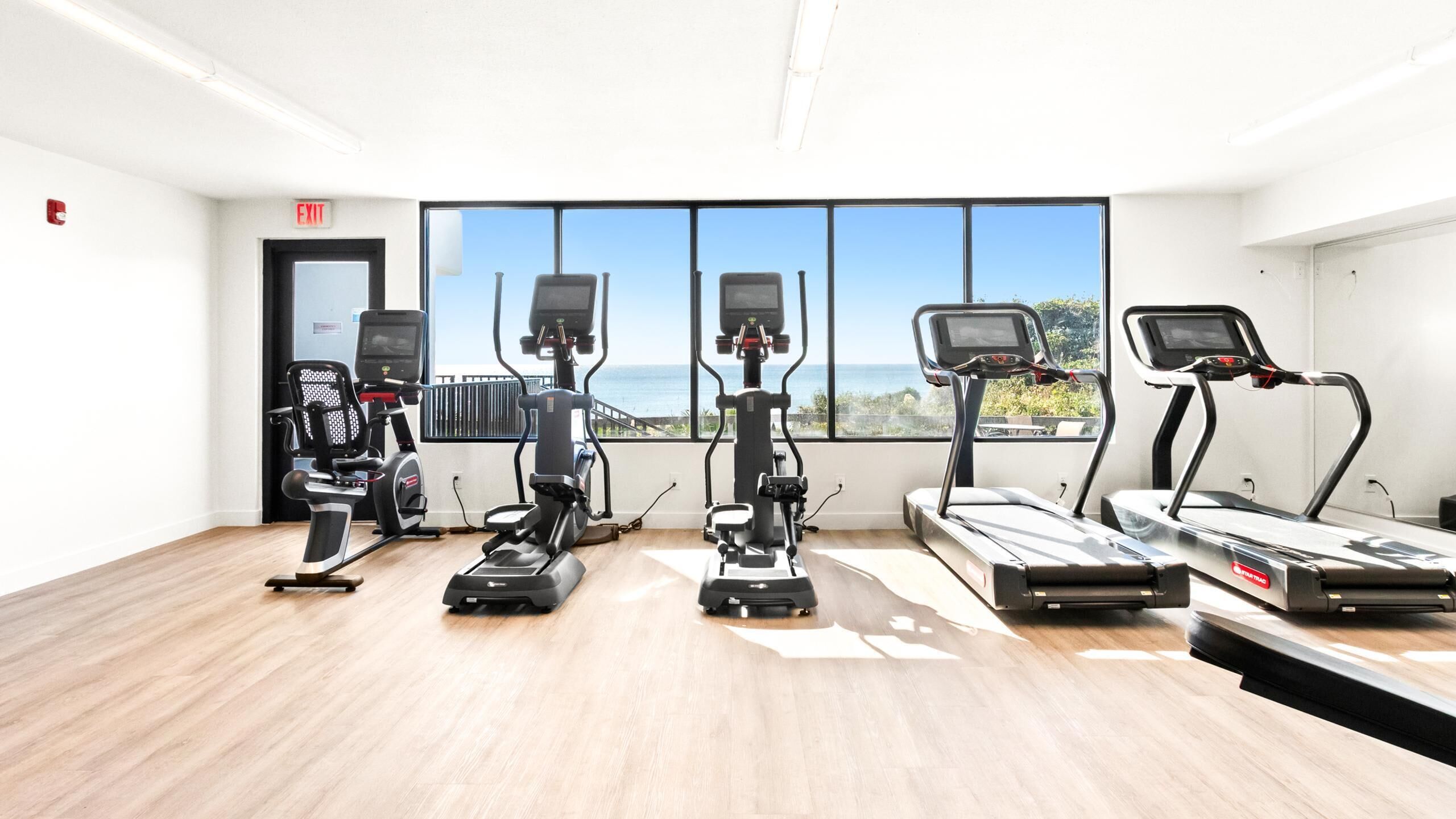 Ein Fitnessstudio mit verschiedenen Cardiogeräten, Holzboden und einem großen Fenster mit Blick auf das Meer.