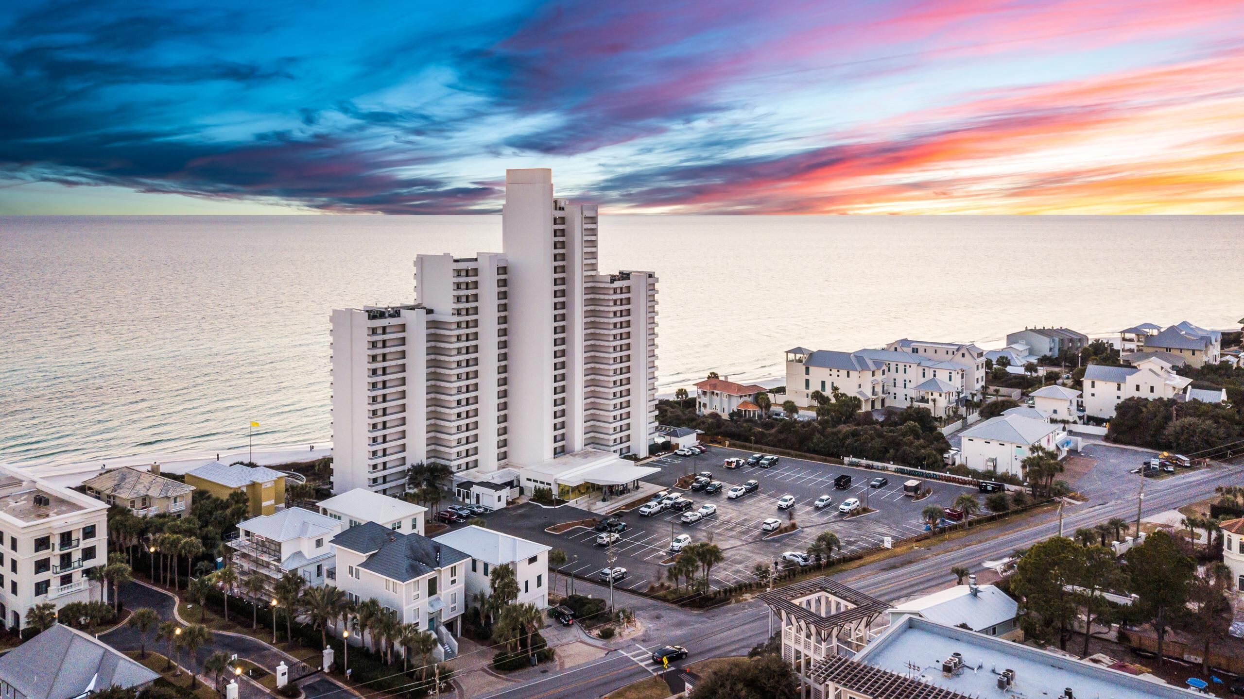 A coastal cityscape with a prominent white high-rise hotel, a parking lot, and a marina.