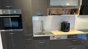 Et moderne køkken med sorte skabe, indbygget ovn, kaffemaskine og en stålvask.