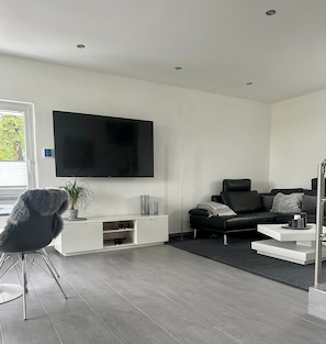 Un salon moderne doté d’un téléviseur à écran plat, d’un canapé en cuir noir, d’un meuble de rangement blanc et d’un fauteuil gris.