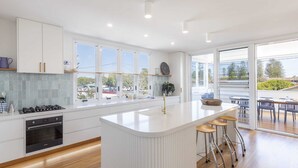A modern kitchen with a central island, white cabinetry, and large windows. - Sandhurst - Oceanstays (Yamba)