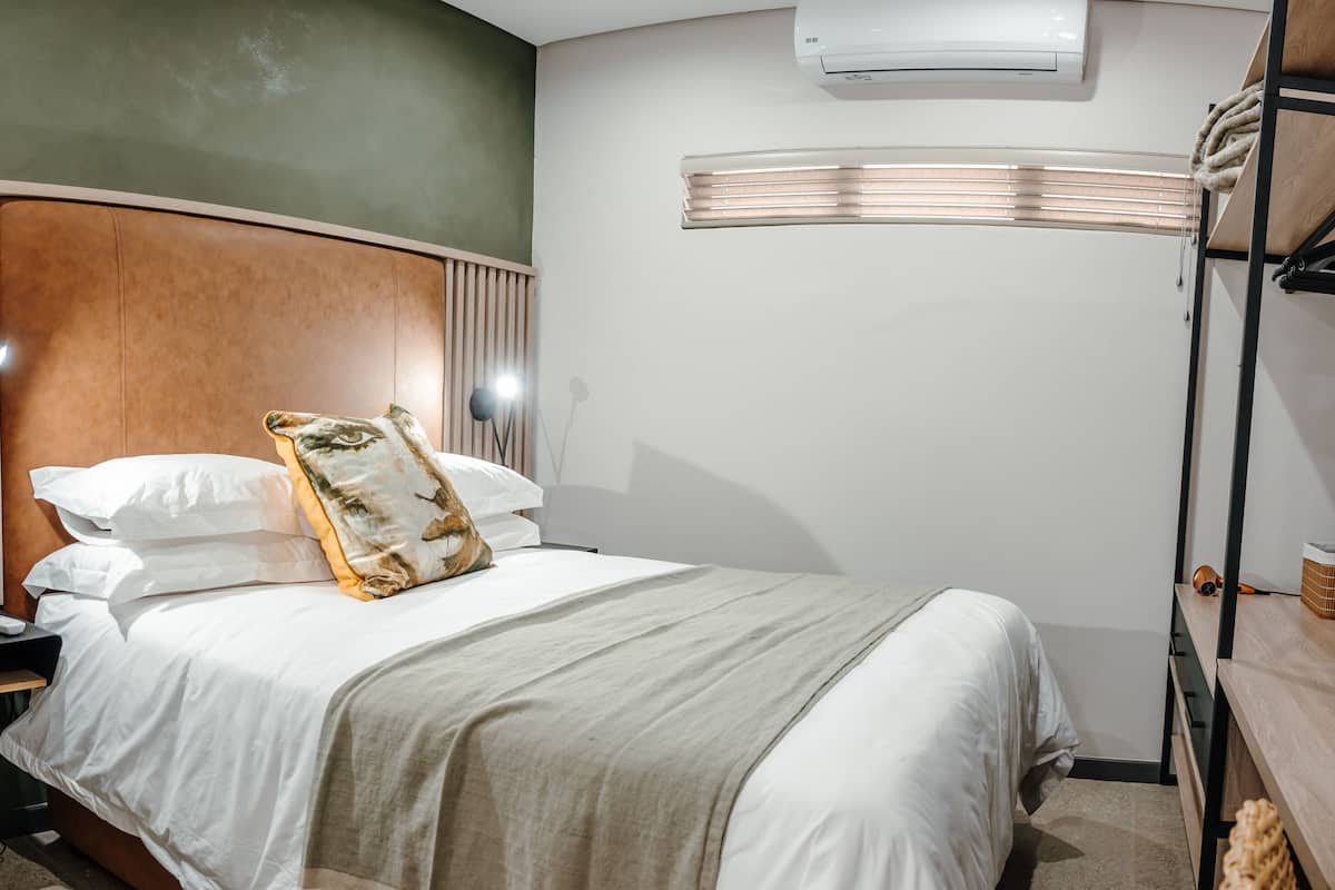 A neatly made bed with white linens and a patterned throw pillow, a bedside lamp, and a wall-mounted air conditioner.