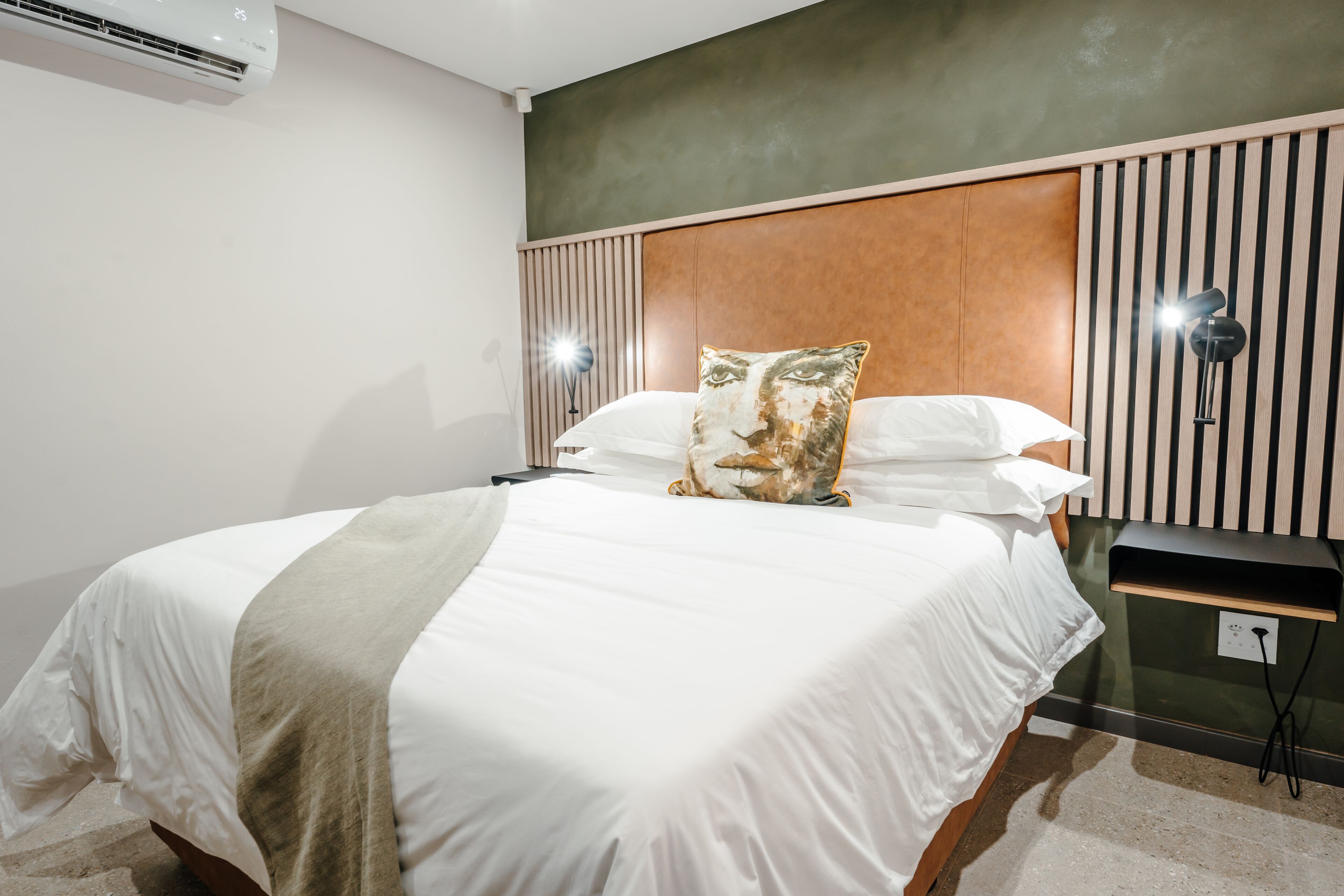A neatly made bed with white linens and a beige throw blanket, a wooden headboard, and a wall-mounted lamp beside it.