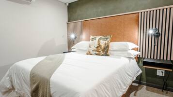 A neatly made bed with white linens and a beige throw blanket, a wooden headboard, and a wall-mounted lamp beside it.