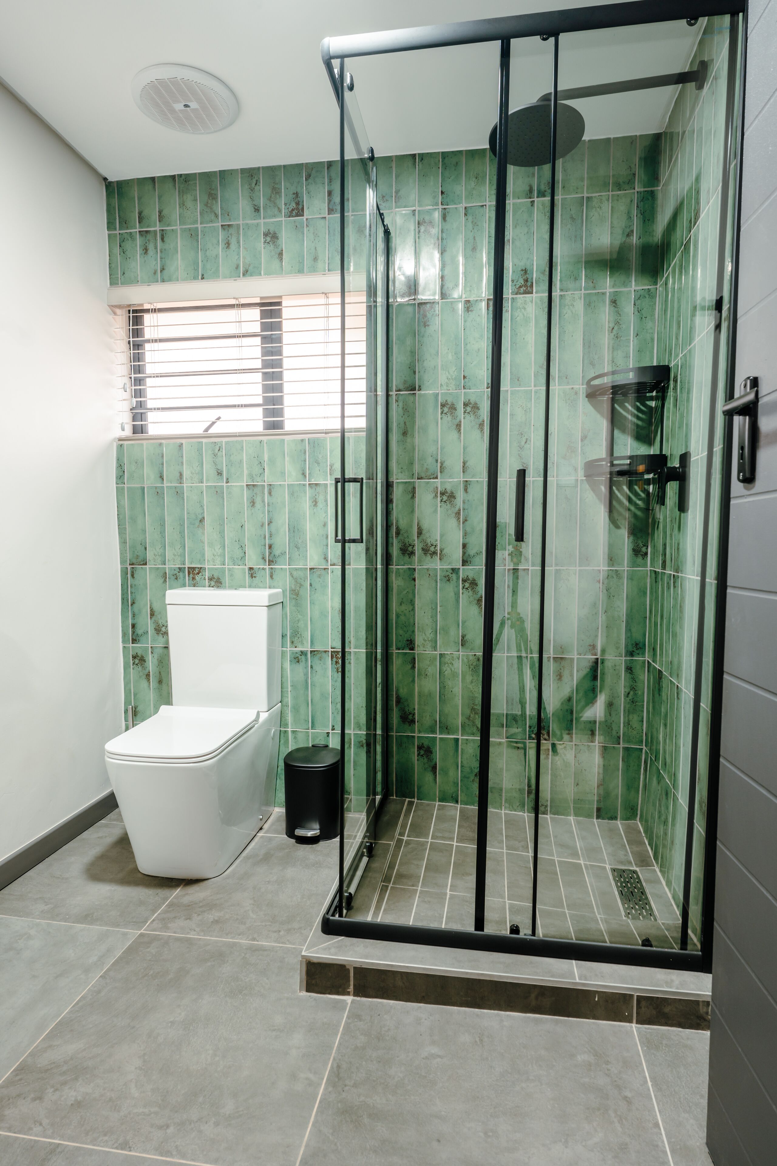 A bathroom with a glass-enclosed shower, a toilet, and a window with blinds.