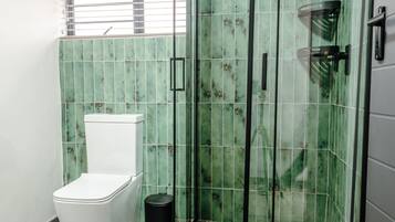 A bathroom with a glass-enclosed shower, a toilet, and a window with blinds.