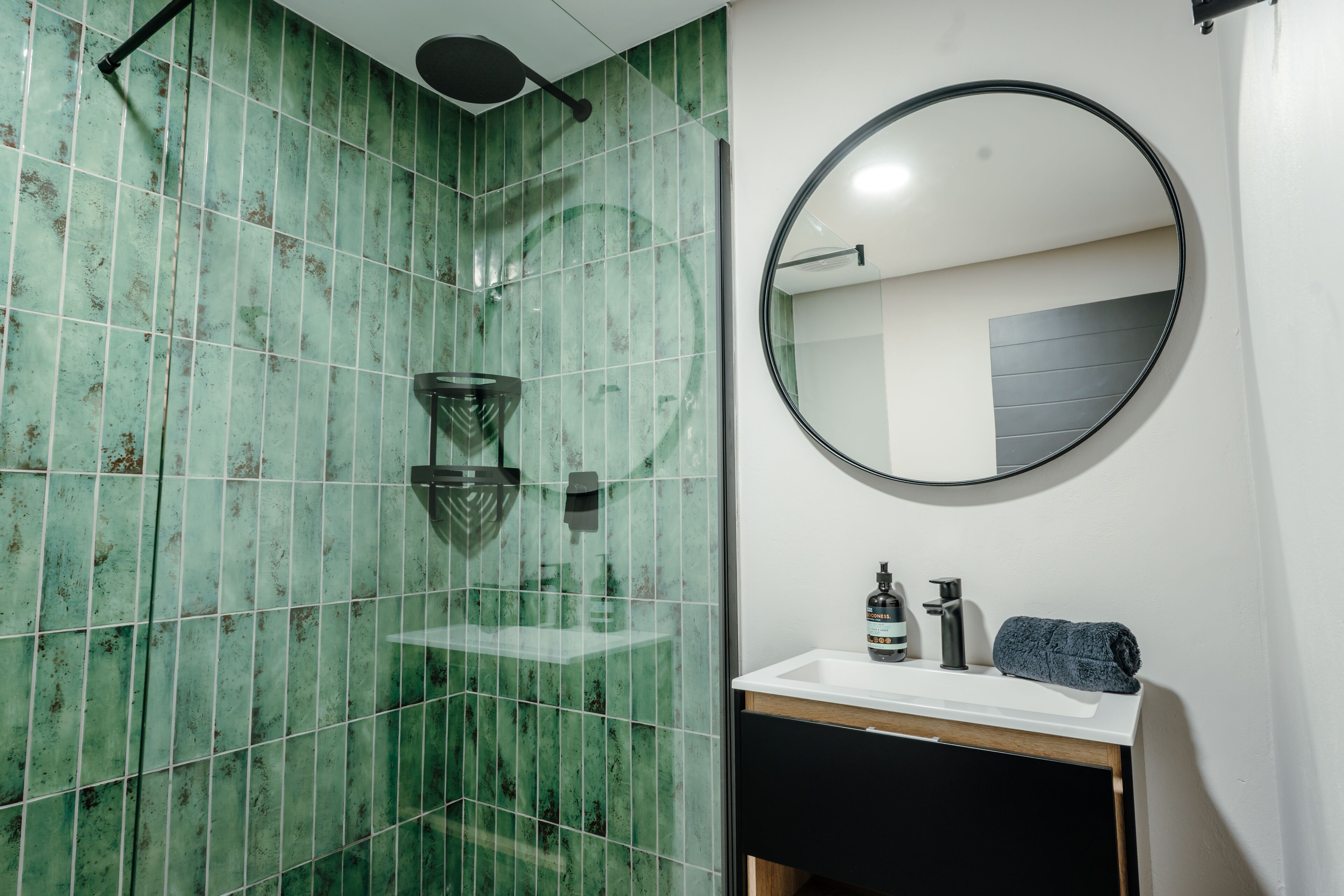A bathroom with green tiles, a glass shower enclosure, a round mirror, a sink with a towel, and a bottle of hand soap.