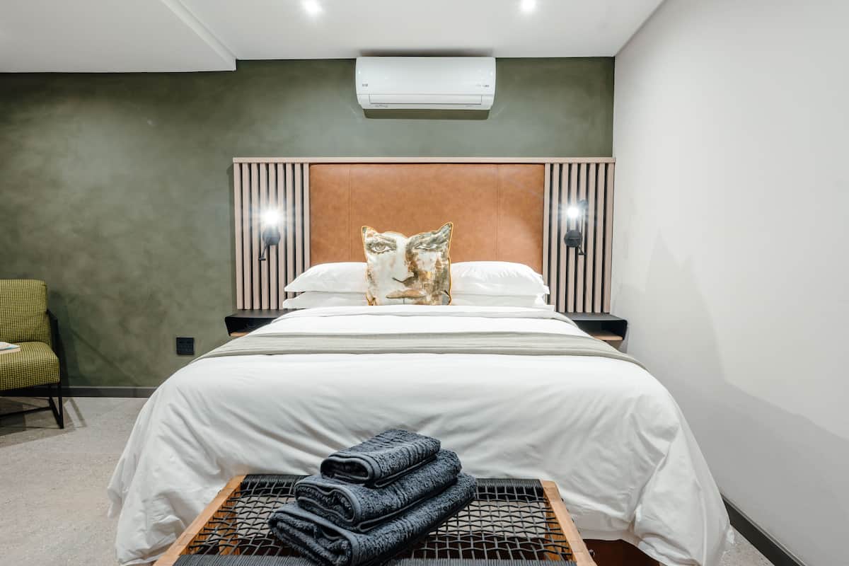 A modern hotel room with a bed, a headboard, a wall-mounted air conditioner, and a small seating area with a chair.