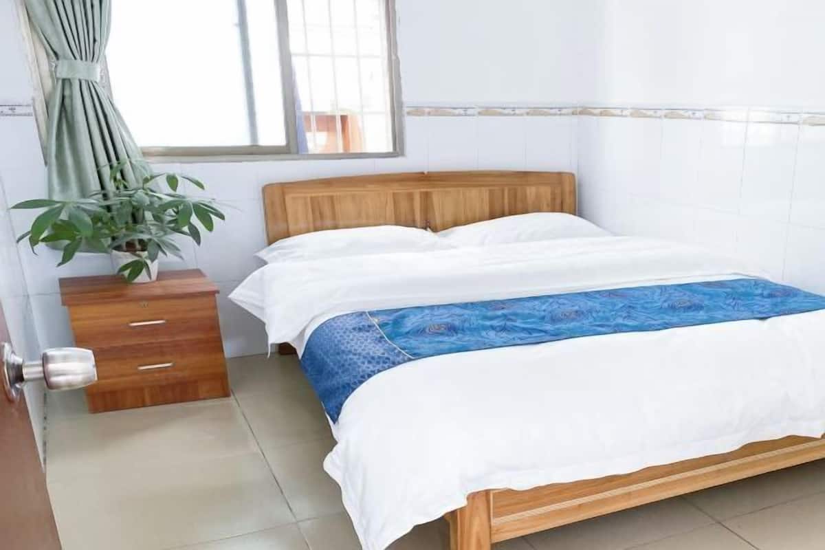 A bedroom with a wooden bed, white bedding, a blue patterned throw, a wooden nightstand, a potted plant, and a window with curtains.