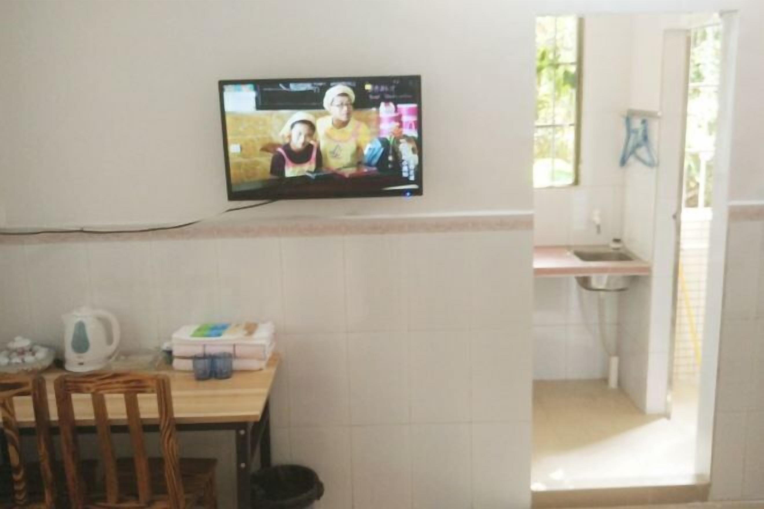 Een kamer met een aan de muur bevestigde televisie, een tafel met stoelen, een keuken met een spoelbak, en een raam.