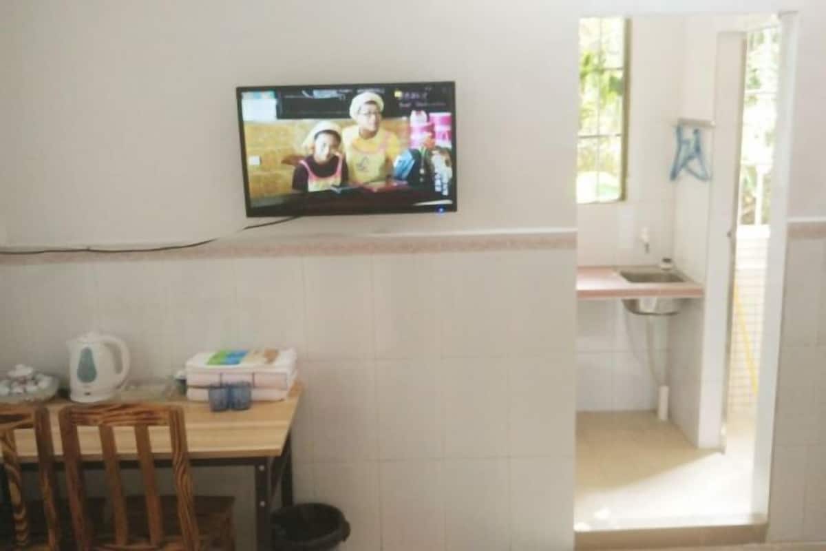 A room with a television mounted on the wall, a table with chairs, a kitchen area with a sink, and a window.