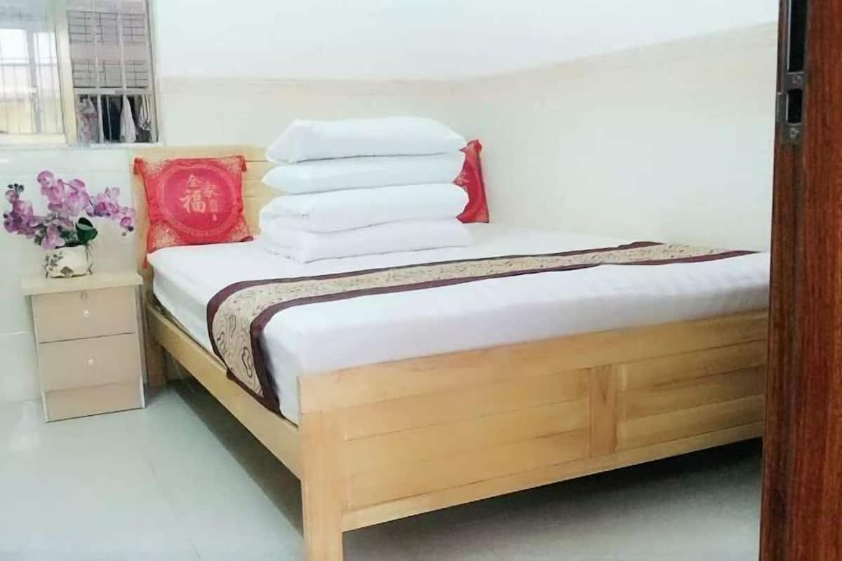 A neatly made bed with white linens and a brown bedspread, a wooden nightstand with a vase of flowers, and a red decorative pillow.