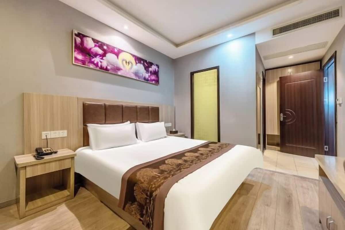 Una habitación de hotel con una cama grande, una mesita de noche con un teléfono, una obra de arte colgada en la pared y una puerta que da a otra habitación.