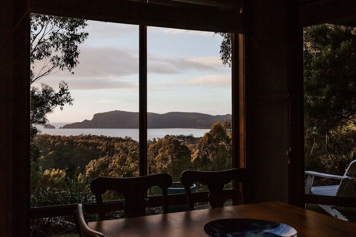 Une pièce dotée d’une grande fenêtre donnant sur un paysage pittoresque, avec des collines et une étendue d’eau. On y aperçoit deux chaises et une table au premier plan.