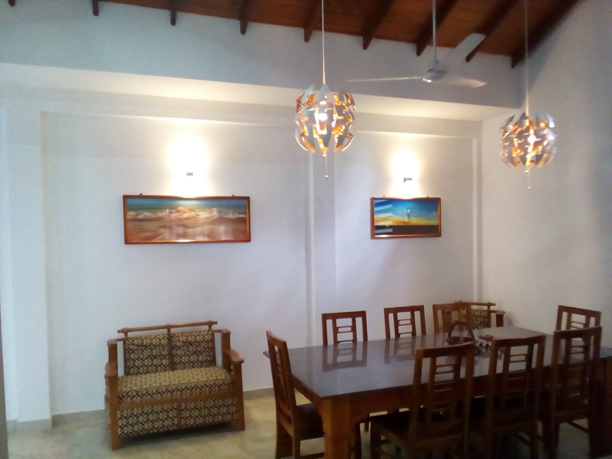 A dining area with a wooden table and chairs, two hanging light fixtures, and two framed paintings on the wall.