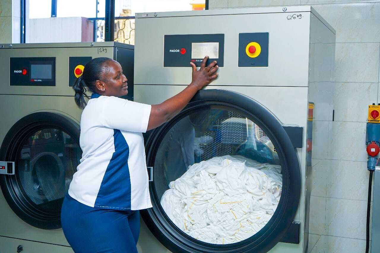 A person operating a large industrial washing machine with a control panel and a large drum filled with white fabric.