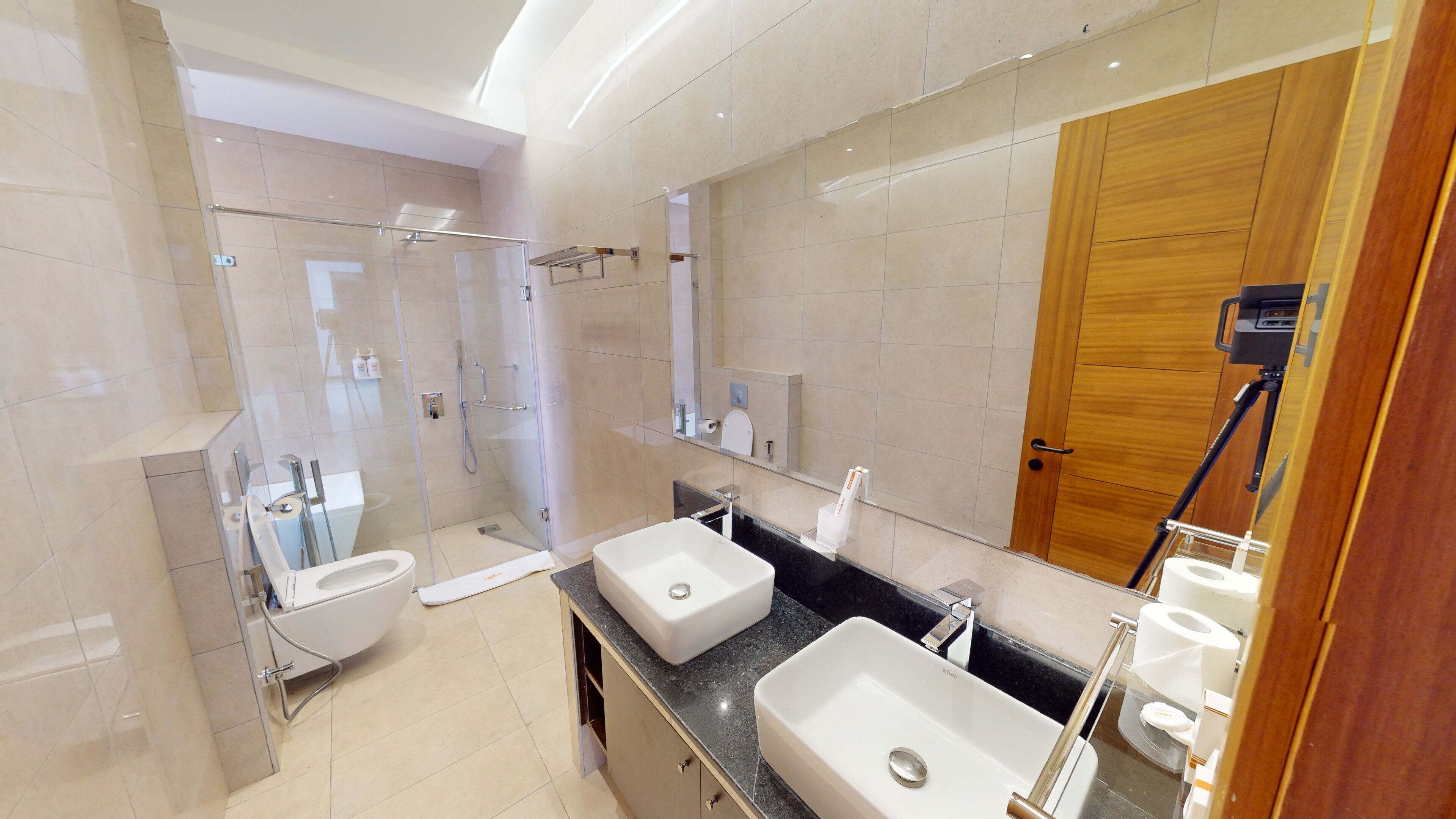 A modern bathroom with a glass-enclosed shower, two sinks, a large mirror, and a wooden door.